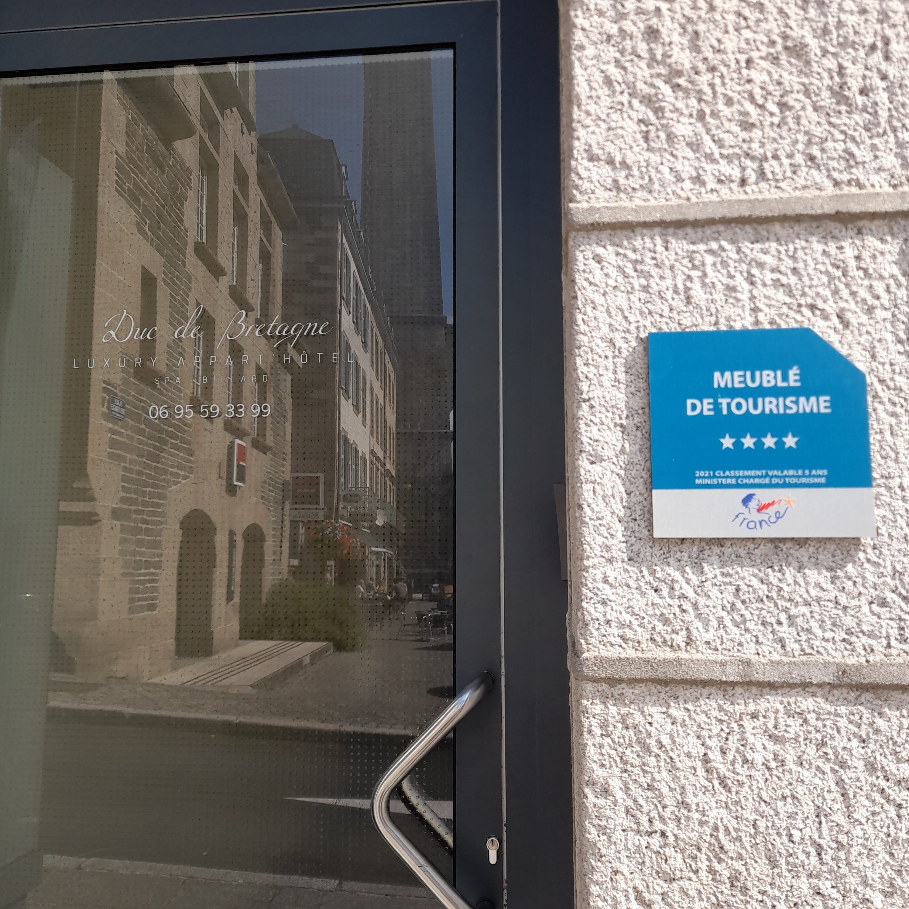 A hotel entrance with a glass door reflecting a street view, a contact number, and a blue sign indicating a tourism award.