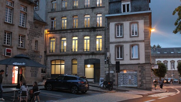 Un immeuble à plusieurs étages avec un café au rez-de-chaussée, un vélo garé à l’extérieur et une rue où circulent des voitures et des piétons.