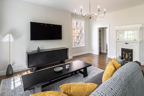 Una sala de estar con televisor de pantalla plana, una mesa de centro de madera oscura, un sofá gris con cojines amarillos, una chimenea blanca y un candelabro.