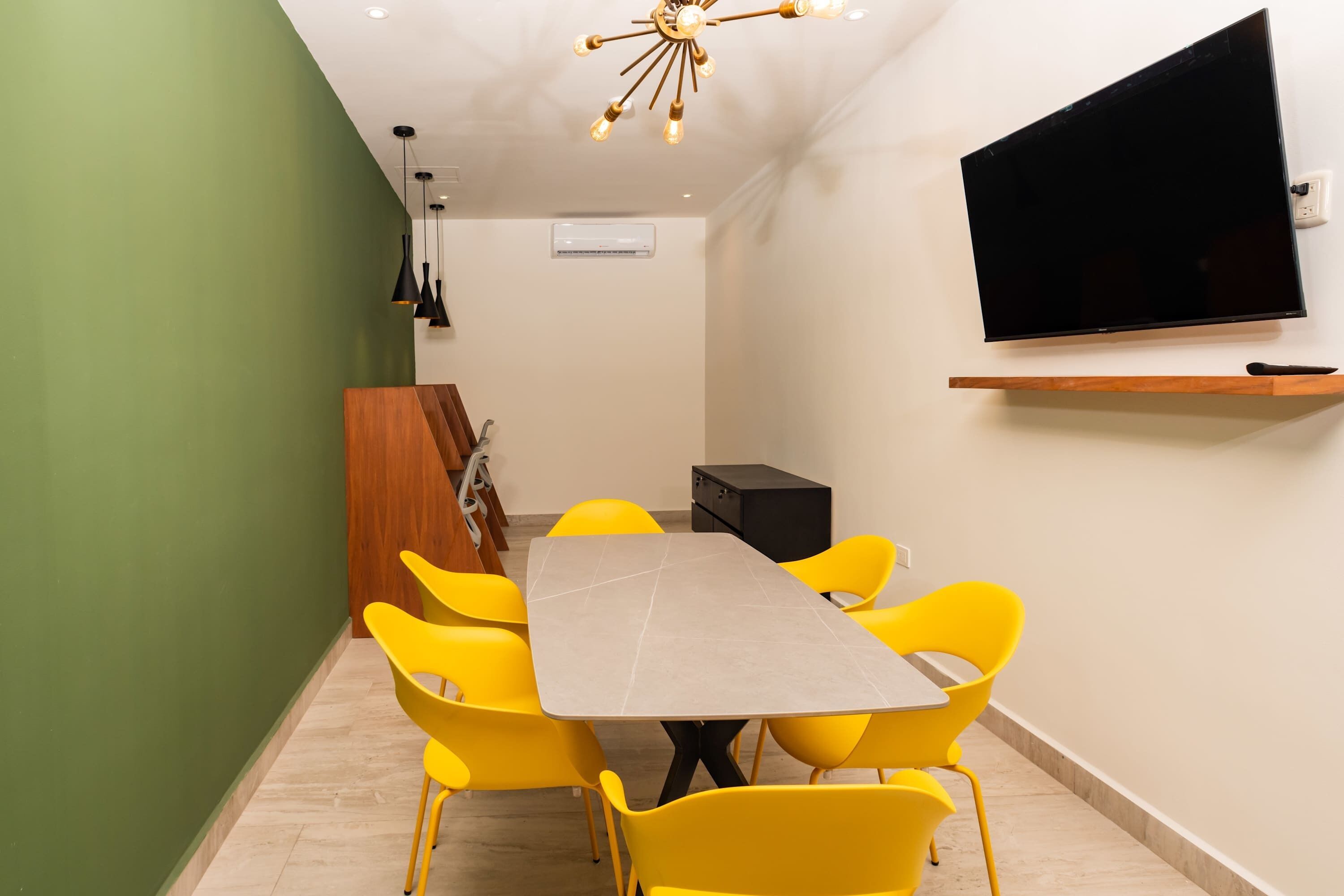 A conference room with a large table, yellow chairs, a flat-screen TV, and a wooden shelf.
