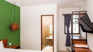 Een hotelkamer met een groene accentmuur, een bed met wit beddengoed, een hangende rieten lamp, een houten deur naar een badkamer en een bureau met een stoel.