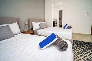 Une chambre d’hôtel avec deux lits, chacun recouvert de draps blancs et agrémenté d’une serviette bleue. On y trouve une table de chevet en bois avec une lampe, ainsi qu’une petite table placée entre les lits. Un tapis à motifs recouvre le sol, et une échelle est visible à l’arrière-plan.