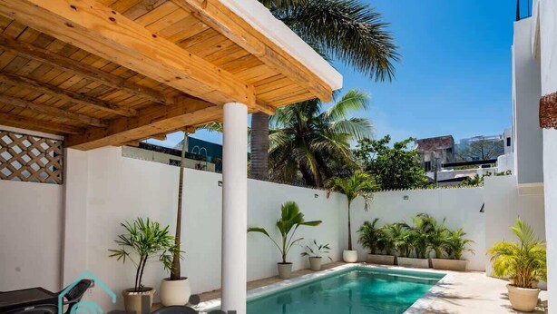 Un área de piscina con un pérgola de madera, plantas en macetas y una piscina azul cristalina.
