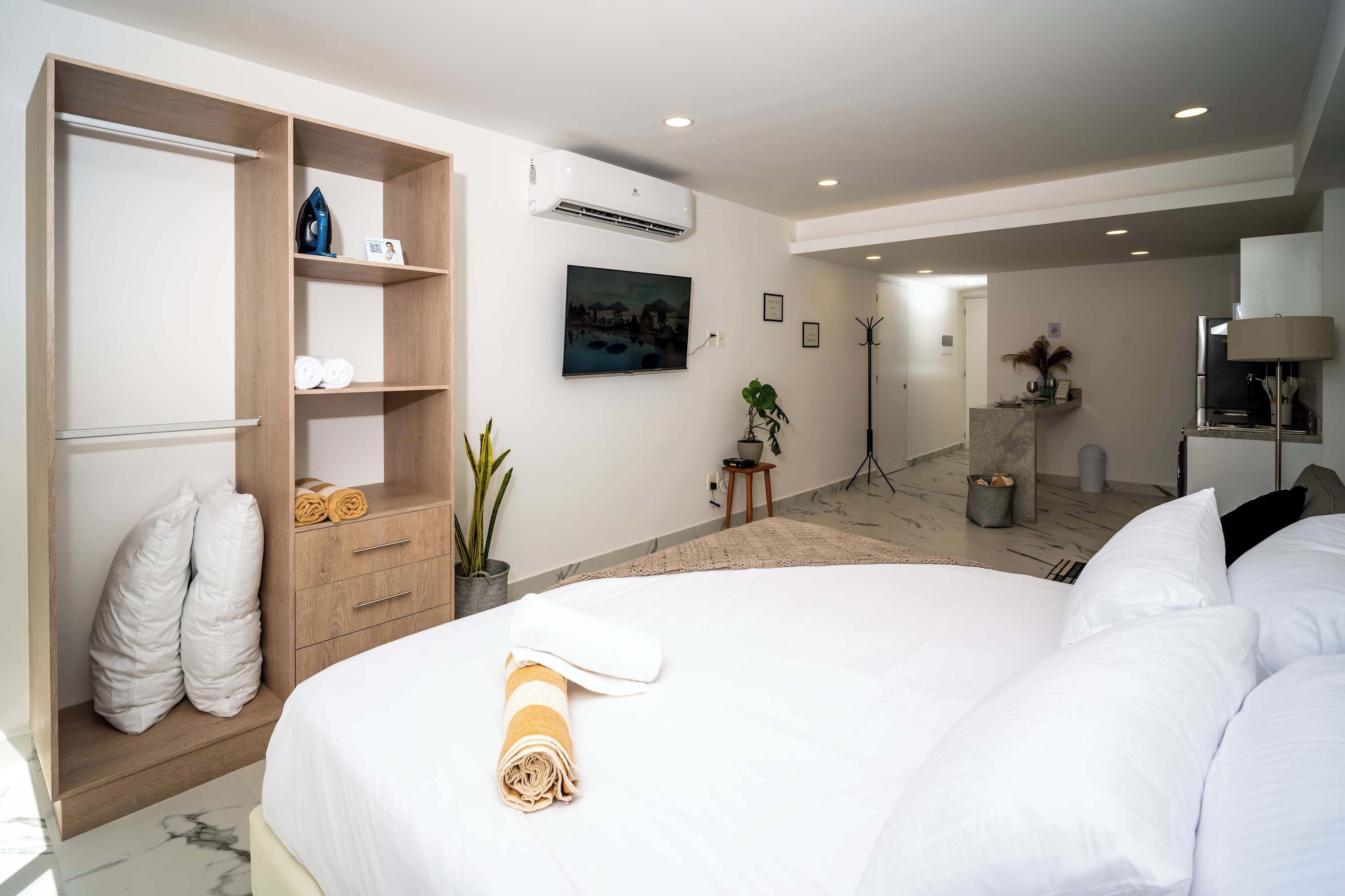 A hotel room with a large bed, a wooden wardrobe, a flat-screen TV, a small table with a plant, and a standing lamp.