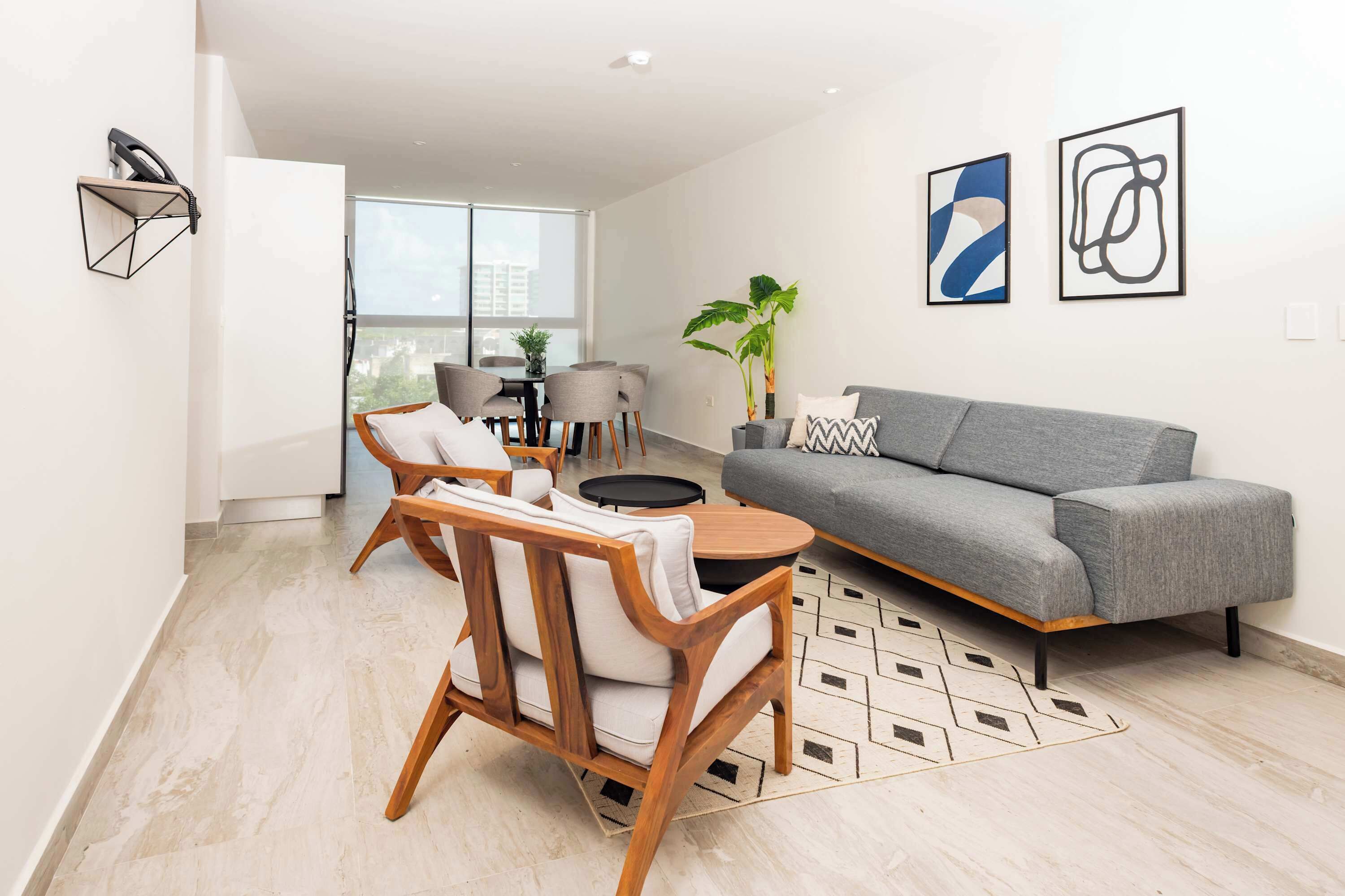 A modern living room with a grey sofa, wooden chairs, a round coffee table, and a dining area with chairs and a table. There are two abstract paintings on the wall and a potted plant.