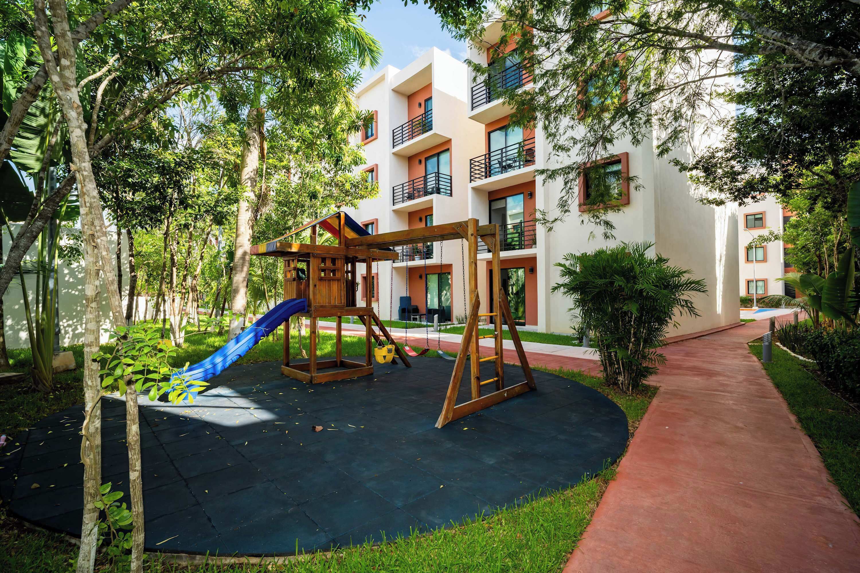Un parque infantil con columpios y un tobogán, rodeado de árboles y un edificio con balcones.