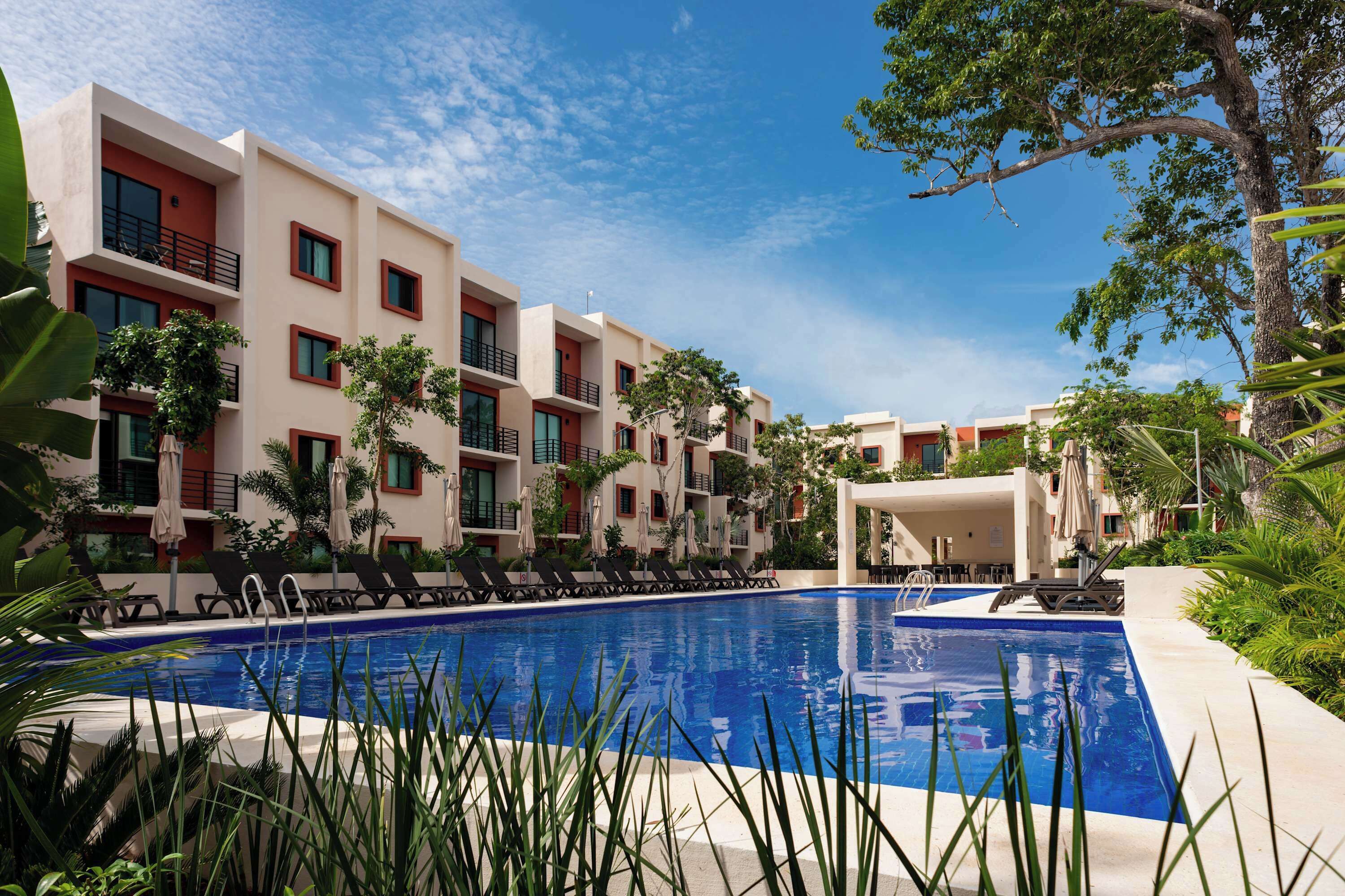 A swimming pool surrounded by modern buildings with balconies, some with red accents, and lush greenery.