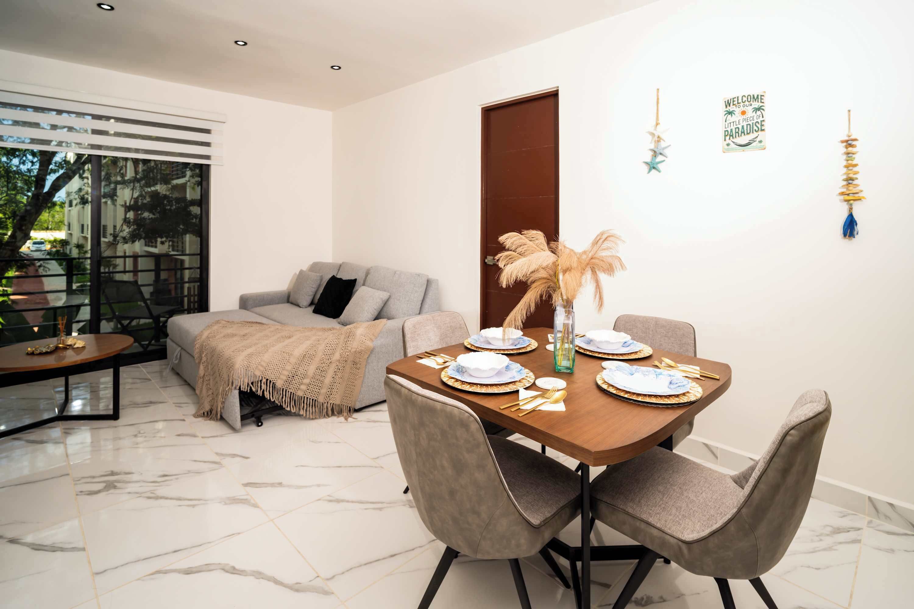 A modern dining area with a wooden table set for four, grey upholstered chairs, and a grey sofa in the background.