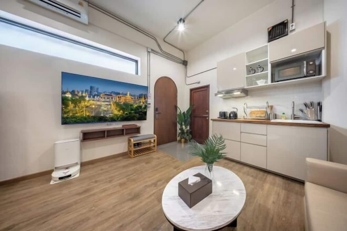 A modern hotel room with a wooden floor, a round marble table, a large cityscape painting, a kitchenette, and a wooden bench.