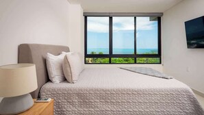 A bedroom with a bed, a nightstand with a lamp, and a large window offering a view of the ocean. - Oceanview with Rooftop Pool in Playa Tortugas (Cancún)