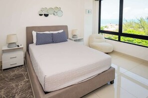 A modern bedroom with a large bed, bedside tables, a chair, and a view of the sea.