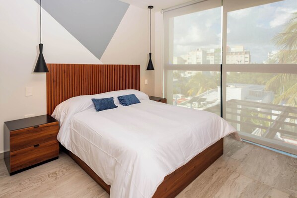 A bedroom with a wooden headboard, a large bed with white bedding, two bedside tables, and a view of buildings through a large window.