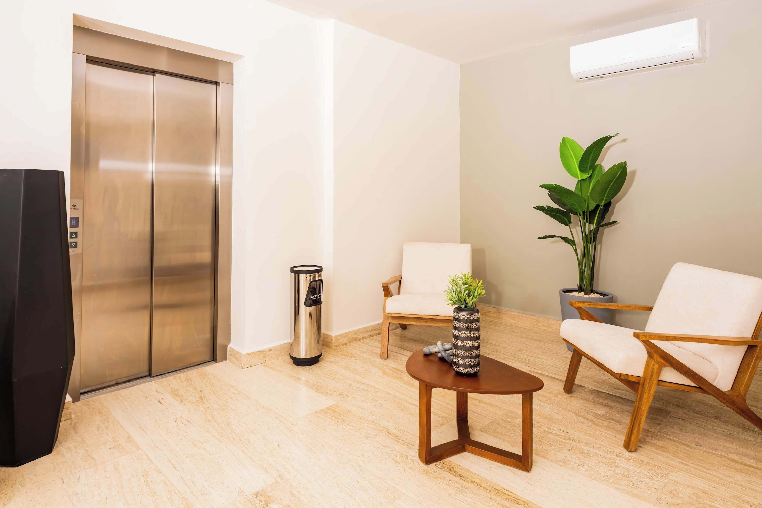 A modern room with a wooden floor, a large metallic elevator, a white chair, a wooden chair, a small round table with a vase, and a potted plant.