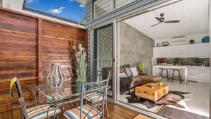 A modern living space with a glass door leading to a kitchen, a wooden deck, and a seating area with striped cushions. - Studio 9 - Luxury Apartment (Byron Bay)