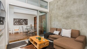 A modern living room with a brown sofa, a wooden coffee table, and a glass dining table with chairs. There is a sliding glass door leading to an outdoor area. - Studio 9 - Luxury Apartment (Byron Bay)