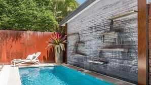 A swimming pool with a brick wall, a palm plant, and a white lounge chair. - Studio 9 - Luxury Apartment (Byron Bay)