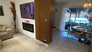 A modern living room with a flat-screen TV, a dining area with a glass table, and a view of the outdoors.