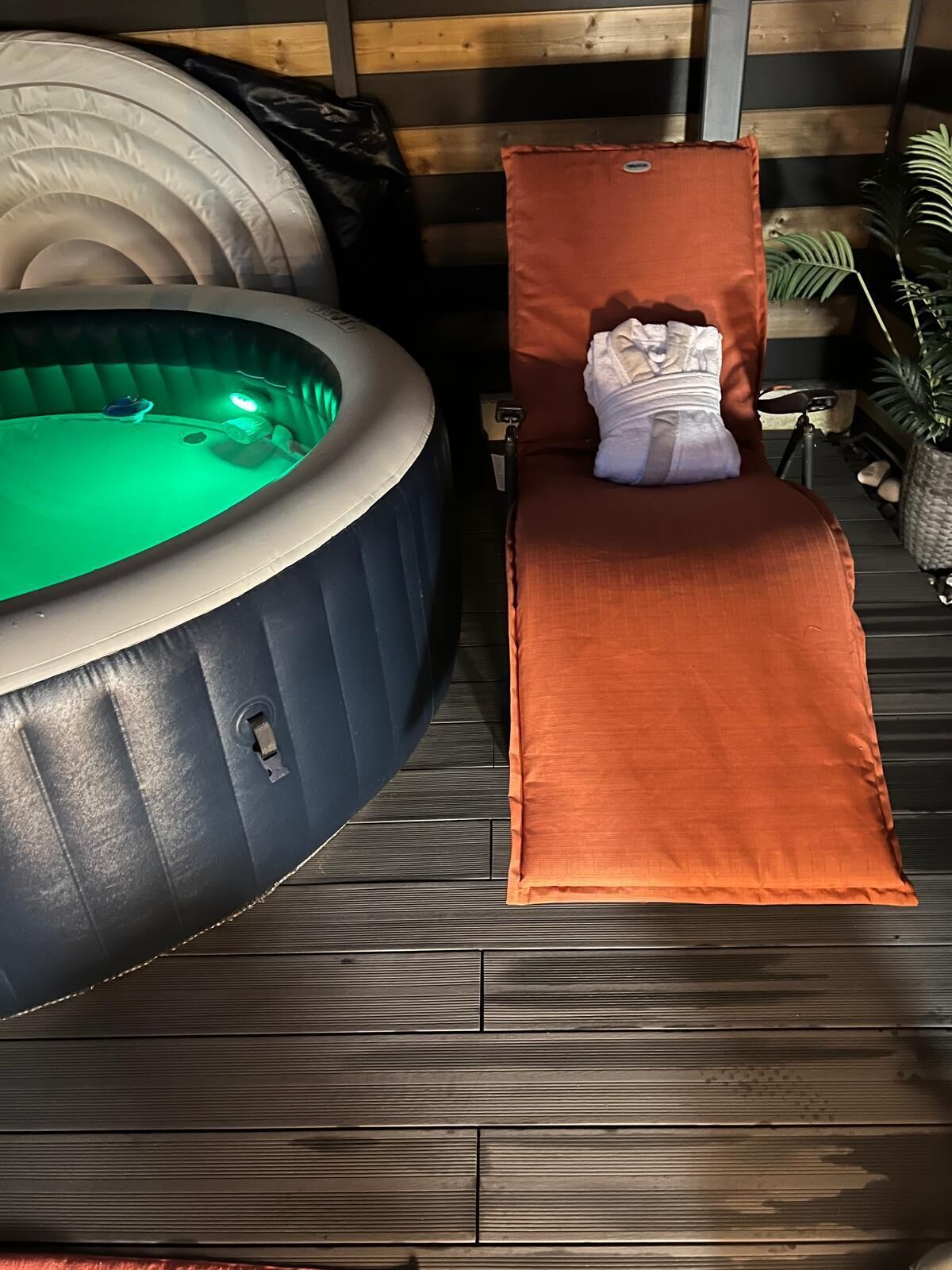 A hot tub with a green light, a brown lounge chair, and a wooden deck.