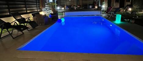 A rectangular swimming pool with a blue tint, surrounded by lounge chairs and a paved deck.