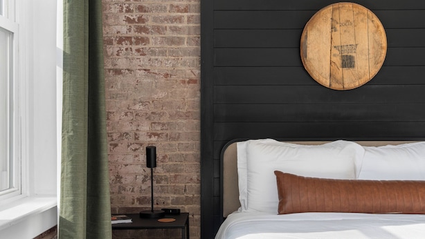 A bedroom with a brick wall, a round wooden headboard, a black nightstand with a lamp, and a bed with white and brown bedding.