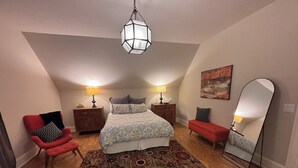 A bedroom with a bed, two bedside tables, a red chair, a large mirror, and a hanging light fixture. - Refined Carriage Apartment in Historic Hope Valley (Durham)
