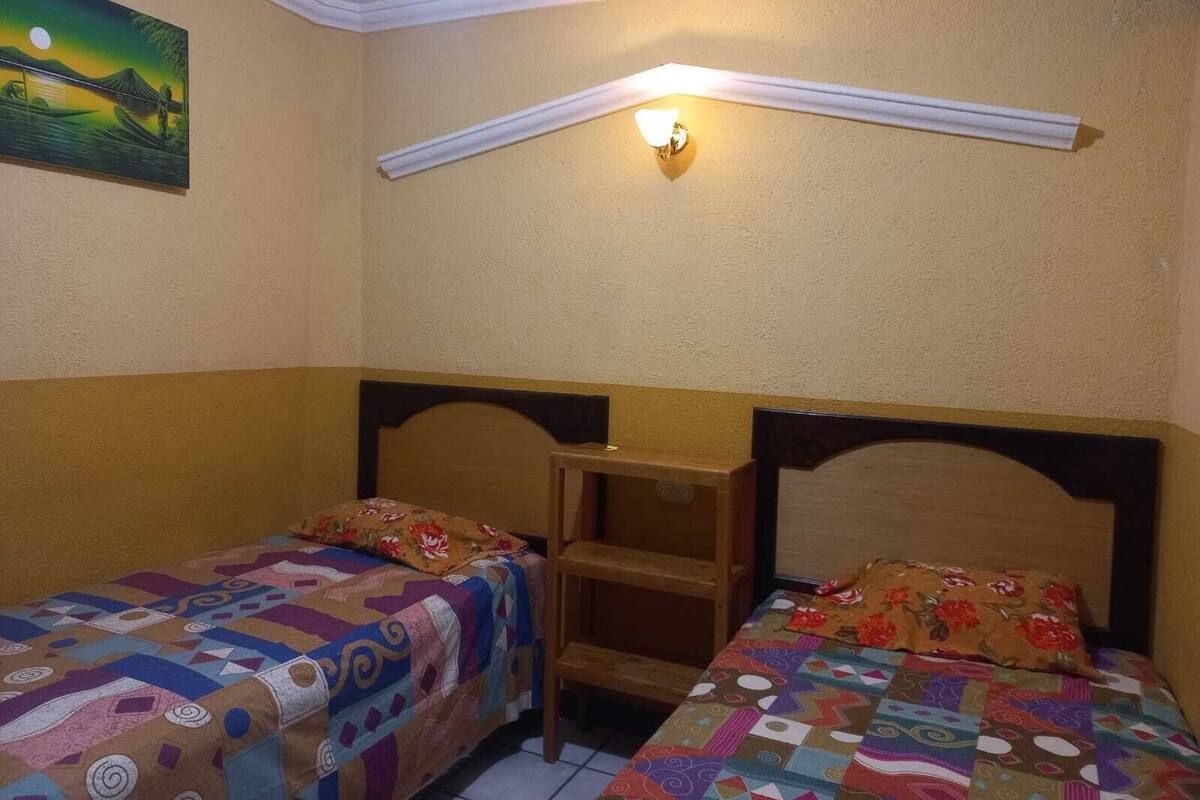 Une chambre avec deux lits, une table de chevet, une lampe fixée au mur et un tableau.