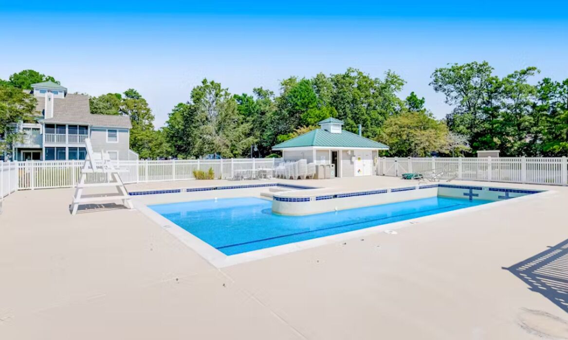 Une piscine rectangulaire avec une terrasse blanche, un petit kiosque et une clôture blanche.