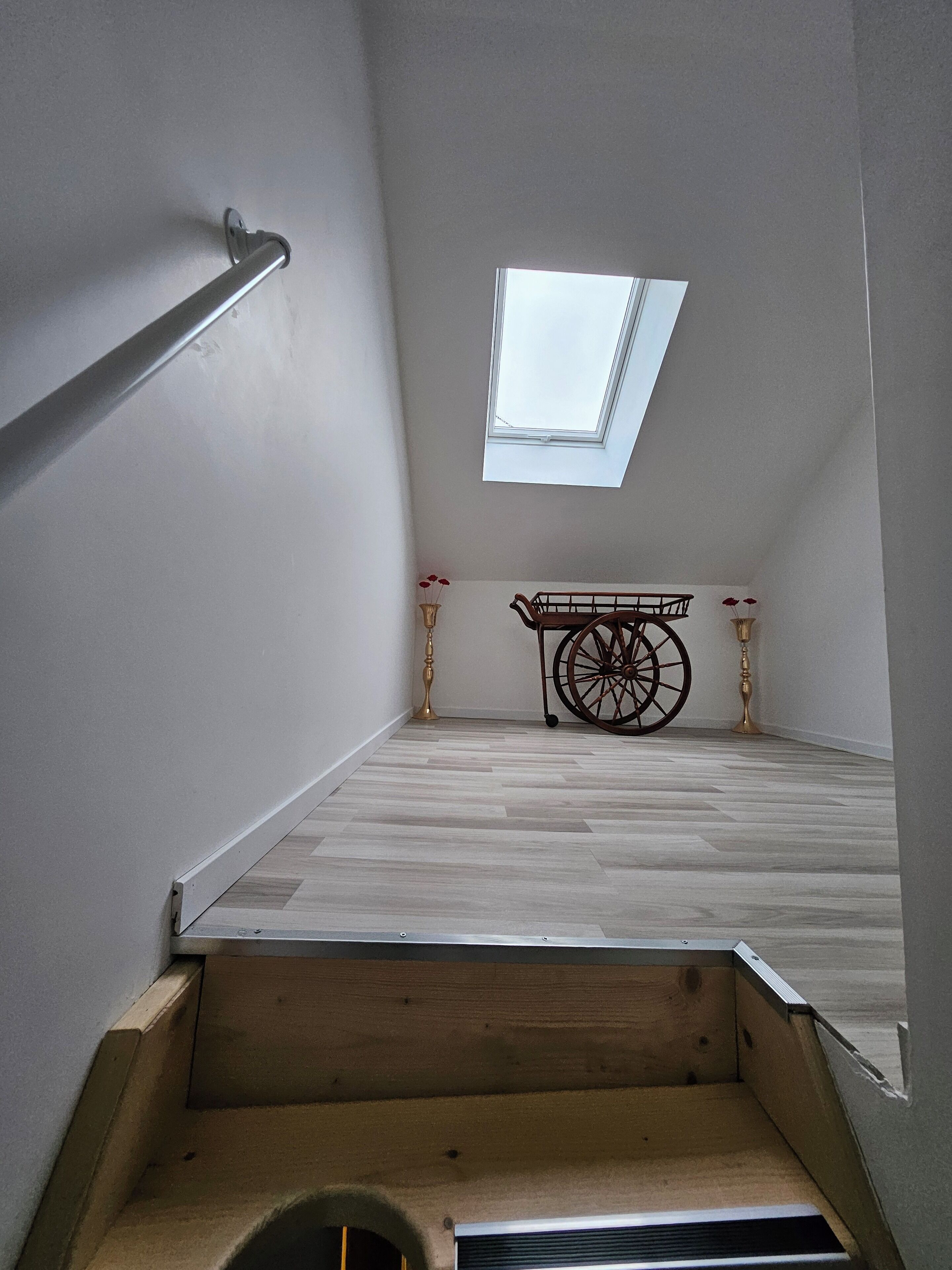 A narrow stairway leading to a room with a skylight, a wooden table with a wheelbarrow, and a vase with red flowers.
