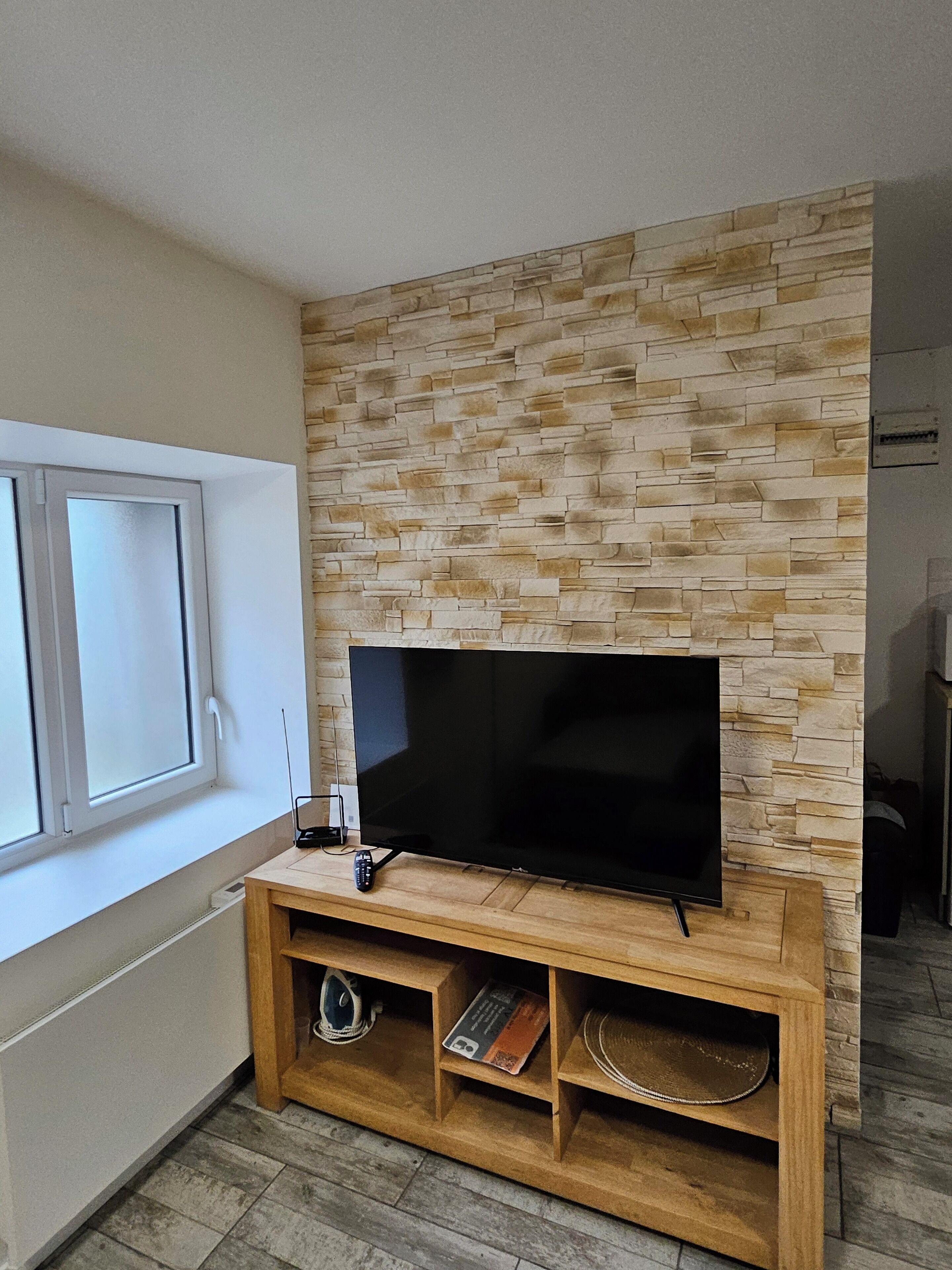 Una sala de estar moderna con un televisor de pantalla plana montado en una pared con diseño de piedra, un mueble de entretenimiento de madera debajo y una ventana a la izquierda.