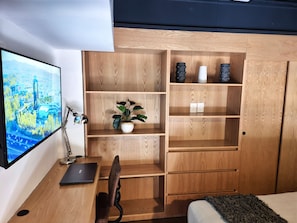 A modern bedroom with a wooden desk, a laptop, a chair, a bed, a TV, and built-in shelves with decorative items. - Lofts La Paz (Guadalajara)