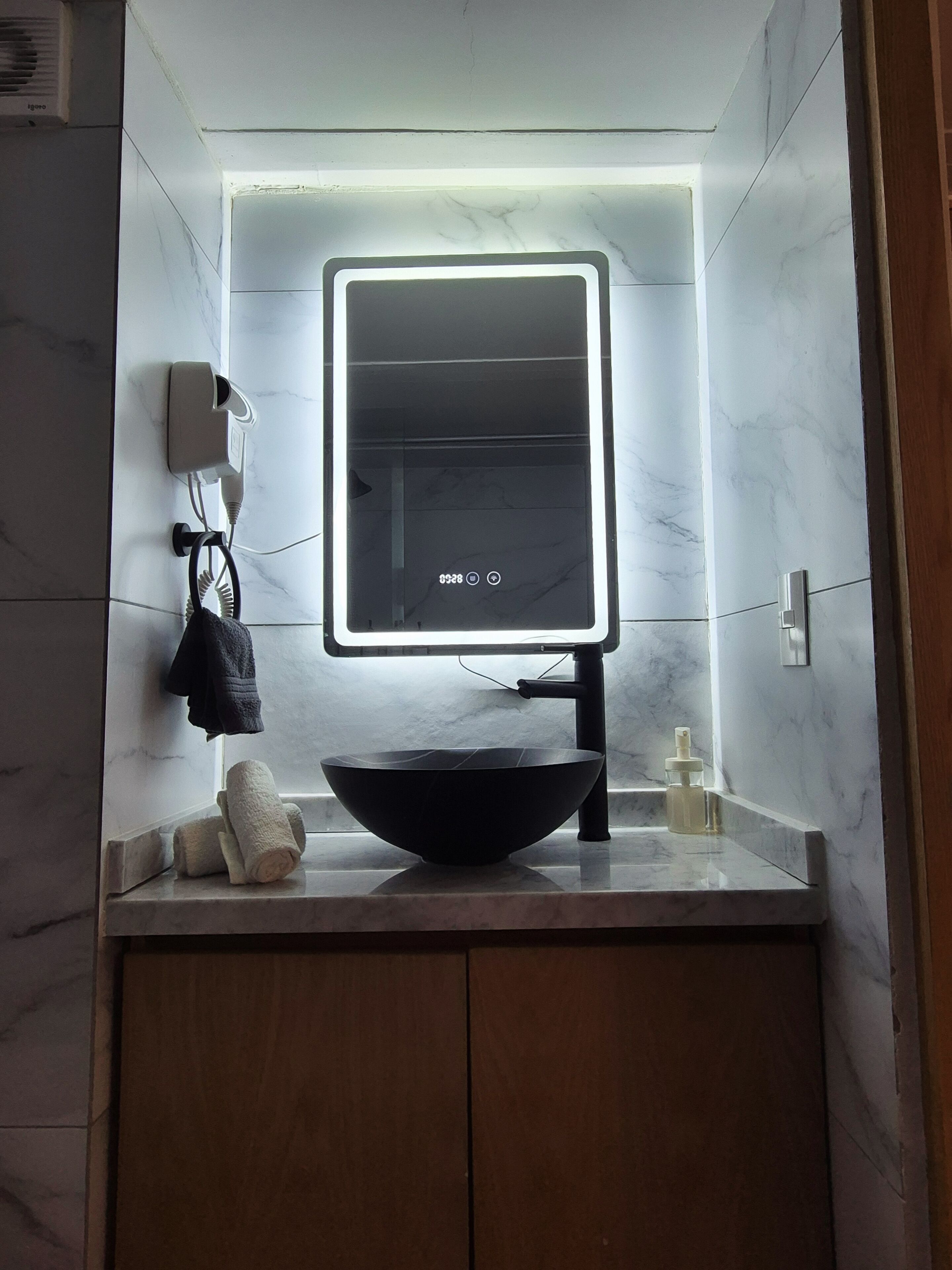 A bathroom with a modern vanity, a black vessel sink, a mirror with LED lighting, a towel rack, and a bottle of hand soap.