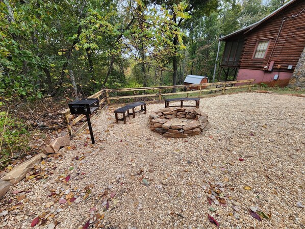 A rustic outdoor area with a stone fire pit, wooden benches, and a wooden fence.