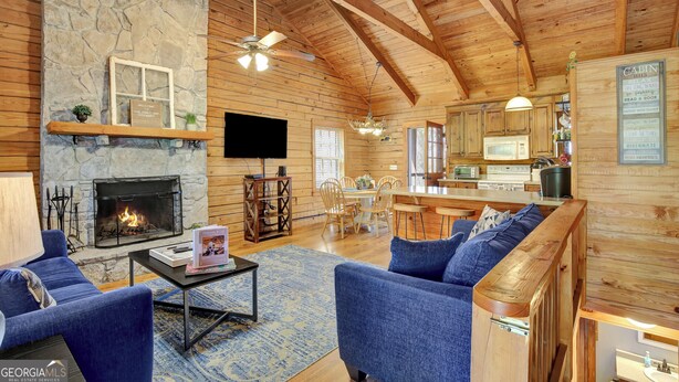A cozy cabin interior with a stone fireplace, blue upholstered seating, a dining area, and a kitchen with wooden cabinets.