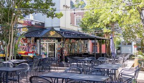 An outdoor cafe with tables and chairs, a storefront with colorful signage, and trees in the background.