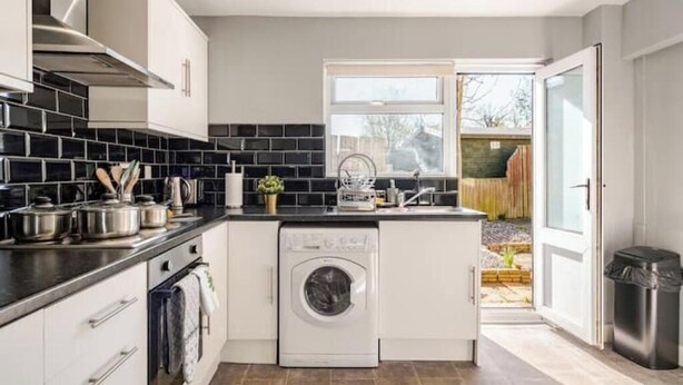 Une cuisine compacte avec des meubles blancs, un dosseret de carreaux noirs, un lave-linge encastré et une fenêtre donnant sur un jardin.