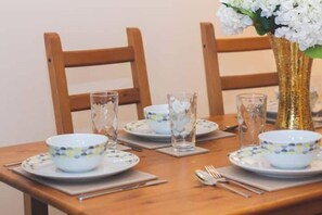 Un tavolo da pranzo apparecchiato con ciotole bianche e bicchieri trasparenti, un vaso dorato con fiori bianchi e sedie in legno.