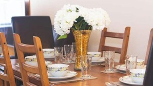 Ein Esstisch, gedeckt mit weißen Tellern, Schalen und klaren Gläsern, mit einer goldenen Vase und weißen Blumen.