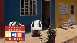 A hostel with two white plastic chairs outside, a door labeled "KIT 02", and a sign indicating "CAMINHITO BRASÍLIA HOSTEL".