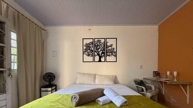 A hotel room with a bed, a fan, a table with glasses, and a wall art piece.