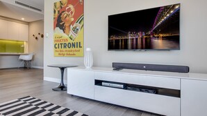A modern living room with a flat-screen TV mounted on the wall, a white entertainment unit, and a poster of a woman holding a bottle. - Spacious 2-bed Apartment Close to City (Sydney)
