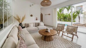 A modern living room with a large window, a round wooden coffee table, a beige sofa, and a few chairs. - Villa Las Casas 2 by BaliSuperHost (Bali)