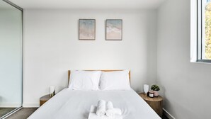 A neatly made bed with white linens and rolled towels, a wooden headboard, two bedside tables with candles and a small plant, and two abstract paintings on the wall. - The Skyline Stopover - Near Sydney Airport (Mascot)