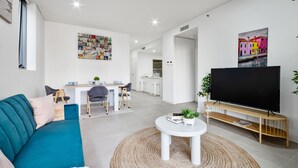 A modern living room with a teal sofa, a flat-screen TV on a wooden stand, a coffee table, and a dining area with a white table and chairs. - The Skyline Stopover - Near Sydney Airport (Mascot)