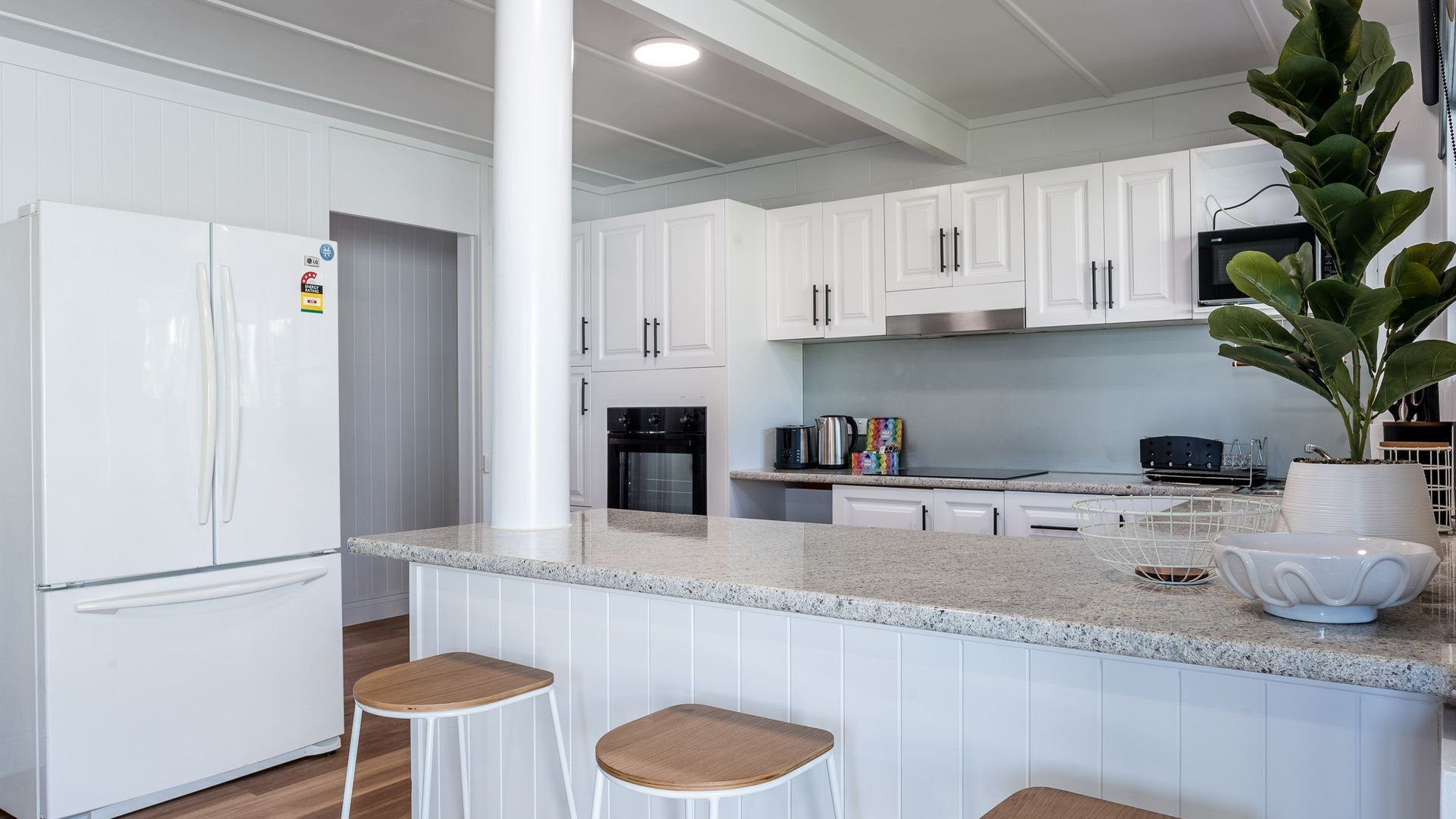 A modern kitchen with white cabinets, a granite countertop, a stainless steel oven, and a microwave. There are two stools and a potted plant on the counter.