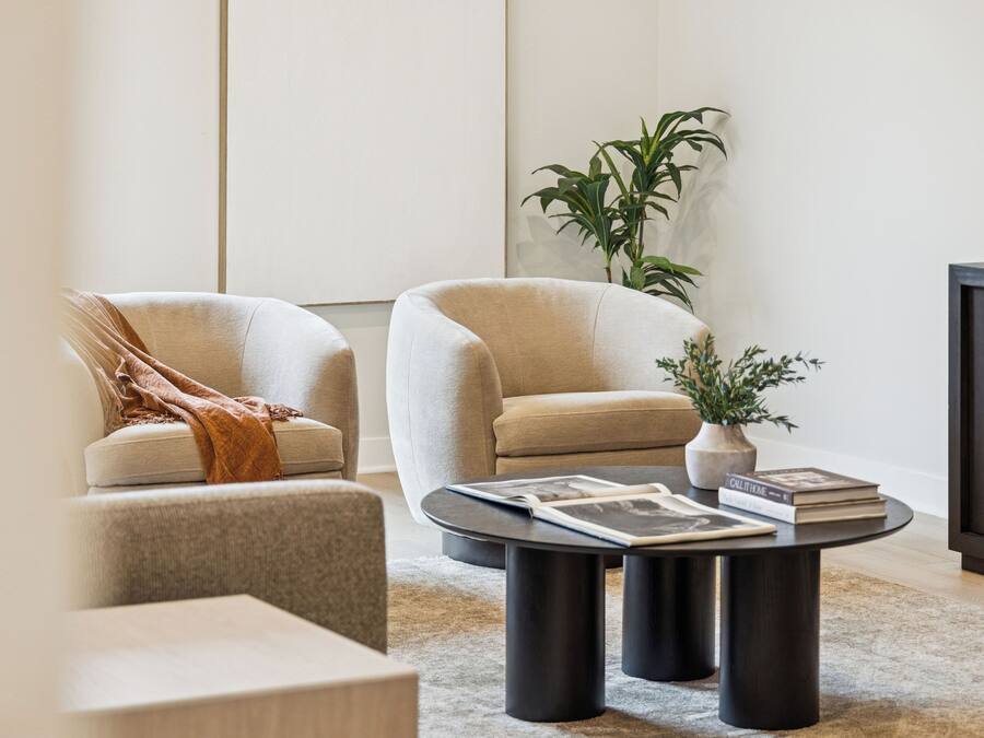 A modern living room with two beige armchairs, a dark coffee table with magazines, a potted plant, and a dark cabinet.