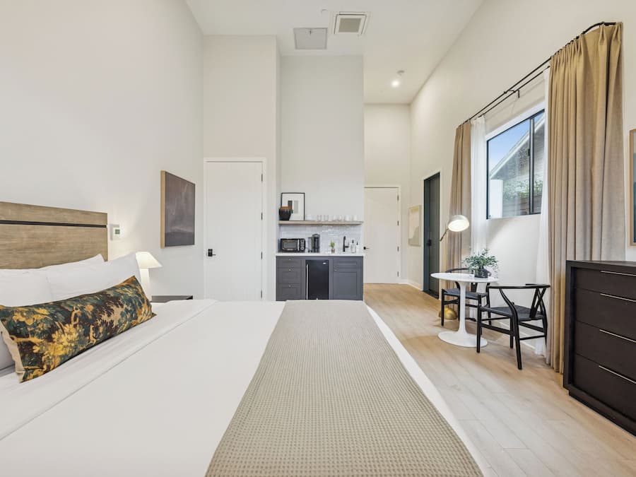 A hotel room with a bed, a desk, a chair, a dresser, a microwave, and a view of the outside.