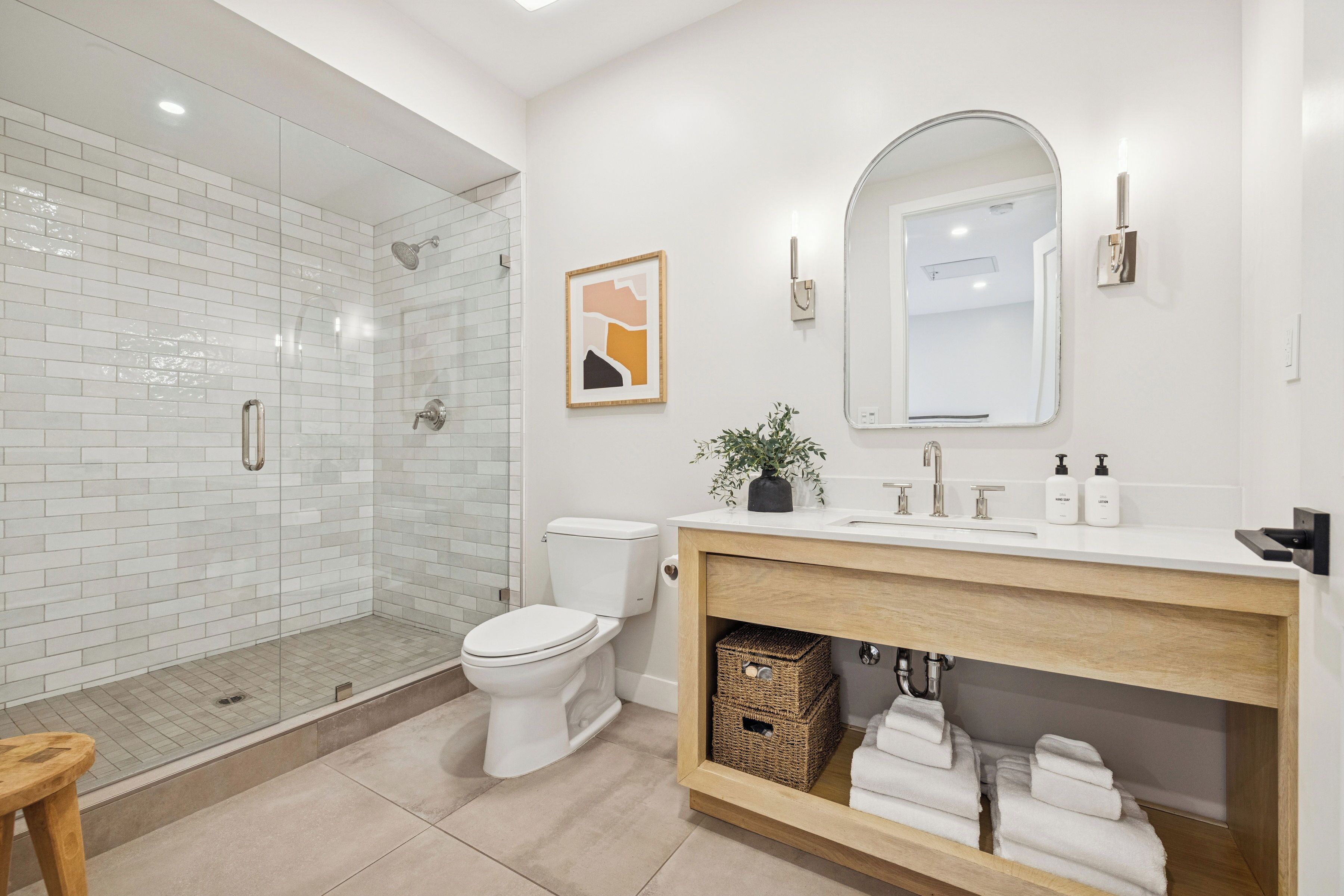 A modern bathroom with a glass shower enclosure, a white toilet, a wooden vanity with a mirror, and a framed artwork on the wall.