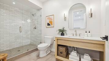 A modern bathroom with a glass shower enclosure, a white toilet, a wooden vanity with a mirror, and a framed artwork on the wall.