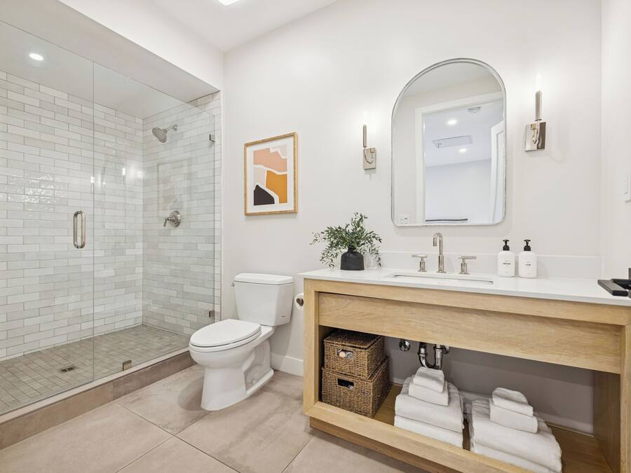 A modern bathroom with a glass shower enclosure, a white toilet, a wooden vanity with a mirror, and a framed artwork on the wall.