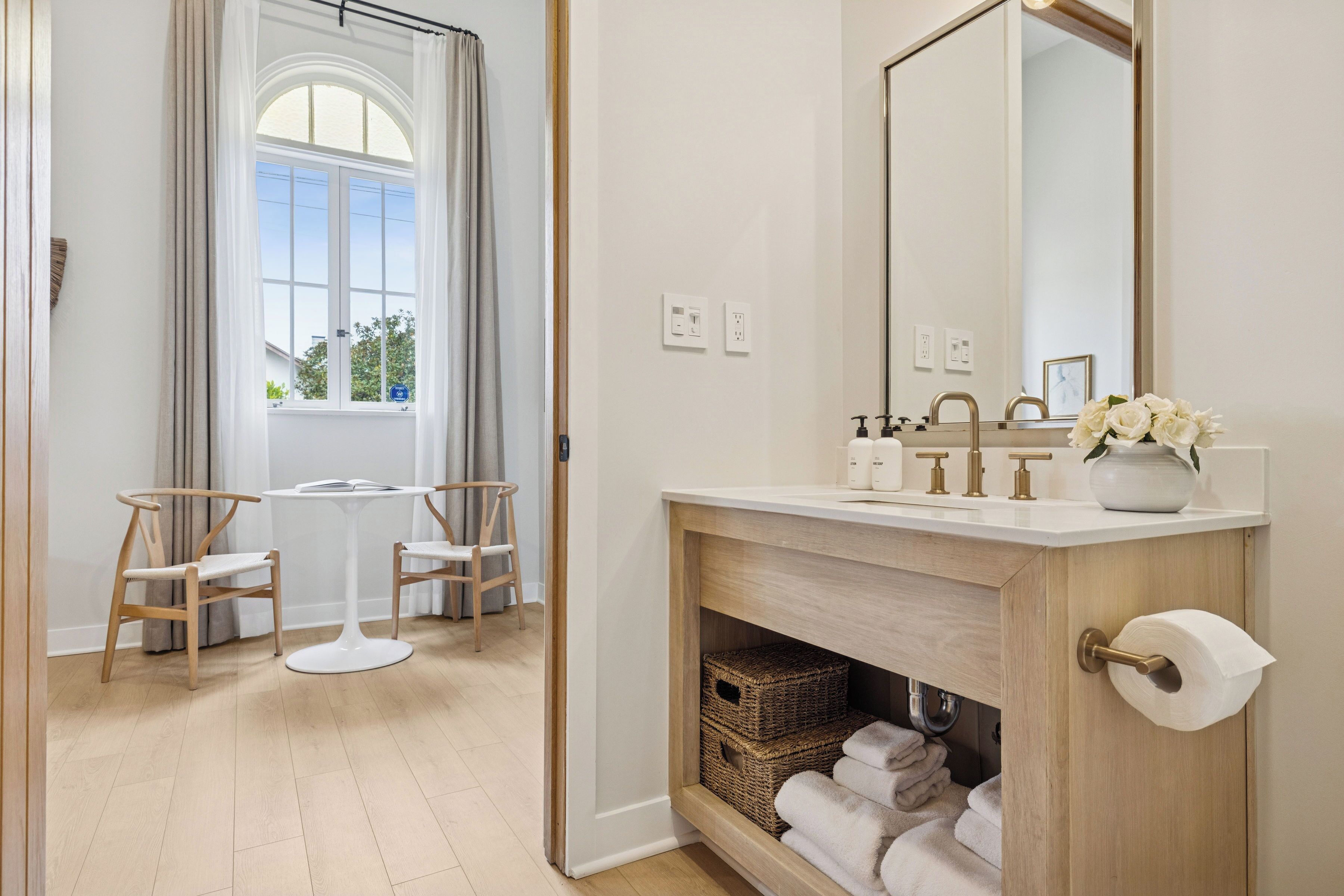 A bathroom with a wooden vanity, a large mirror, and a window with curtains. There are two wooden chairs and a small table.